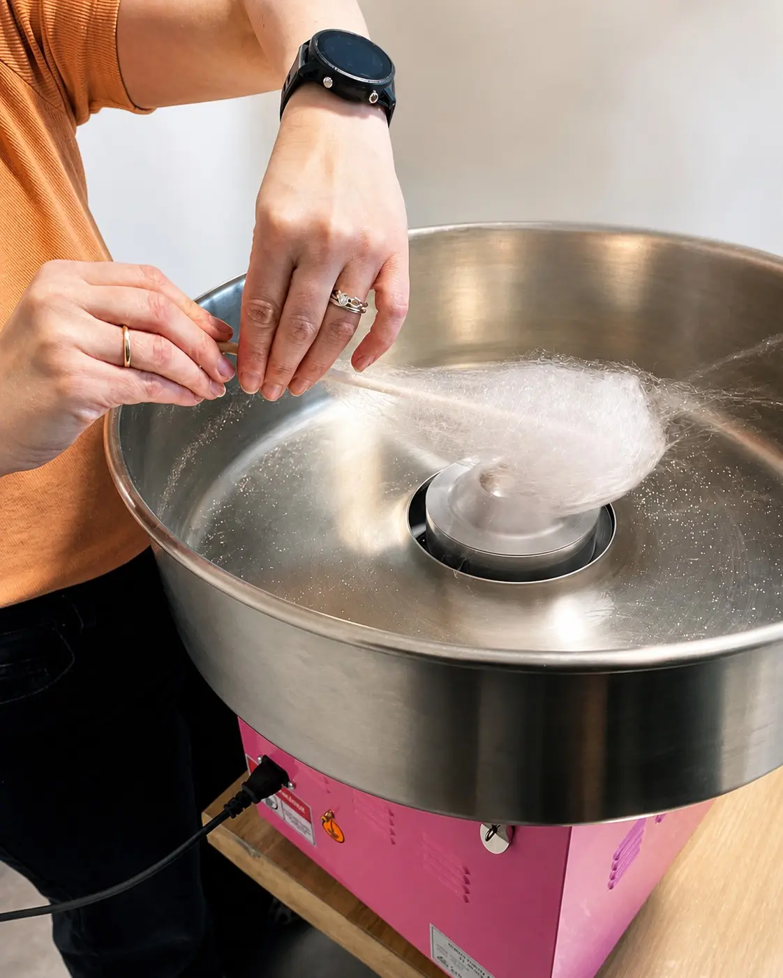 Adult hands spinning pale pink candy floss inside the machine bowl.
