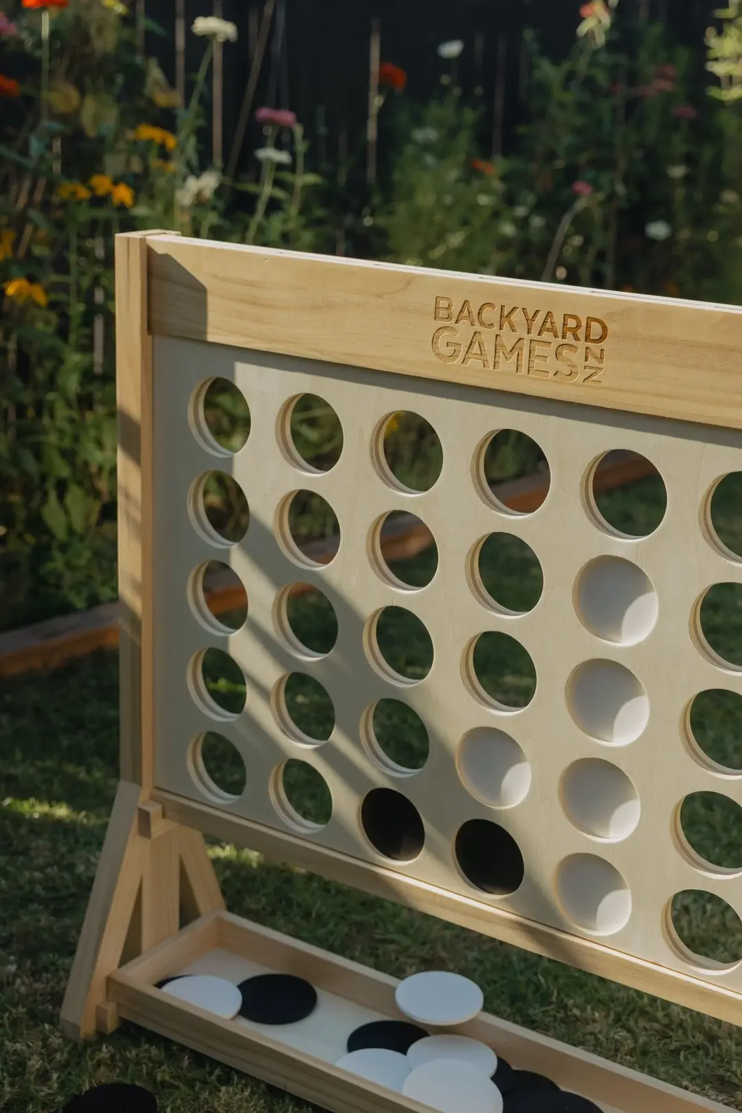 A giant wooden Connect 4 frame with black and white discs, standing on grass in front of a flower garden in afternoon sun.