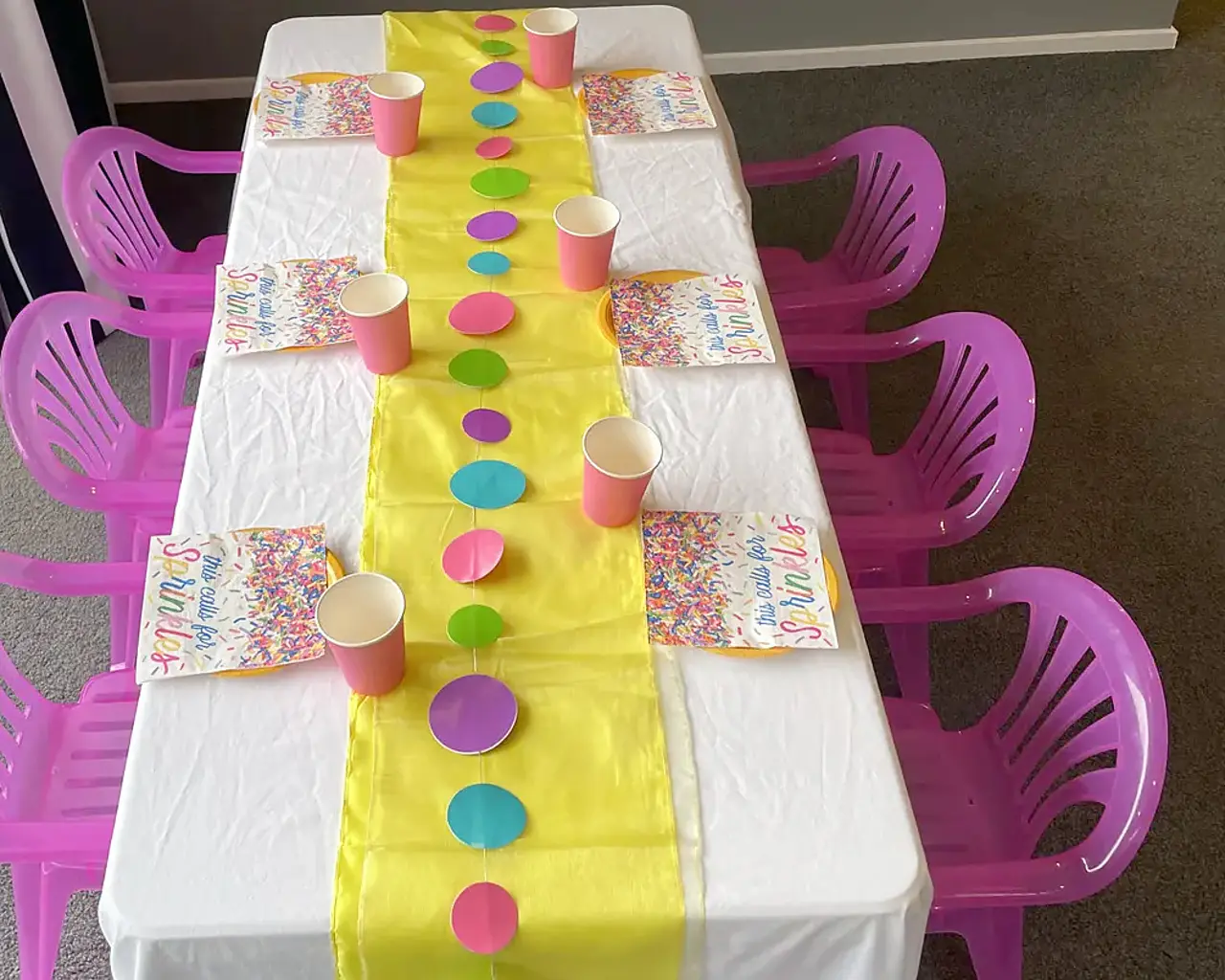 A Playday party table set for six children: a white-clothed table with a yellow satin runner, coral paper cups, sprinkle-print plates and a paper disc garland, with bright pink children’s chairs down both sides.